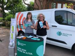 Petra vom Digitalen Engel und Kooperationspartnerin Frau Andermann stehen am Info-Stand vor dem Info-Mobil und halten zwei Flyer. Sie lächeln in die Kamera.