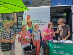 Besucher:innen stehen vor dem Infomobil und Johannes zeigt einer Dame etwas auf dem Smartphone