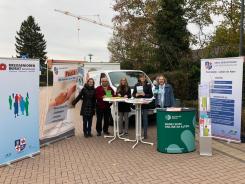 Fünf Personen stehen vor dem Info-Mobil und halten Flyer in die Kamera. Um sie herum stehen drei verschiedene Roll-Ups.