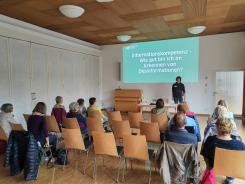 Die Teilnehmenden sitzen auf Stühlen im Raum und blicken zur Wand nach Vorne