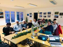 Gruppenbild der Teilnehmenden. Acht Damen und Herren sitzen an einem winterlich geschmücktem Tisch. Im Vordergrund sind ein Laptop und ein Beamer zu sehen.