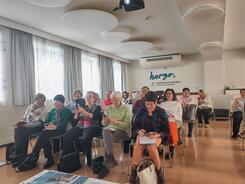 Die Teilnehmenden sitzen auf Stühlen im Raum und schauen zur Präsentation an der Leinwand