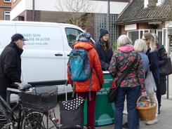 Pera steht in Winterjacke und Mütze vor dem Info-Mobil am Info-Stand. Um den Stand herum stehen fünf Damen mit Einkaufstaschen und ein Herr mit einem Fahrrad.