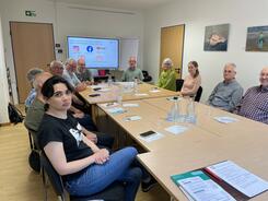 Die 11 Teilnehmenden aus Wunstorf sitzen an einem rechteckigen Gruppentisch und lächeln in die Kamera. Hinter ihnen ist die Endfolie der Präsentation.