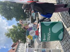 Theresa und eine Besucherin unterhalten sich am Infostand. Hinter ihnen sind ein Brunnen sowie Häuser und zwei Bäume zu sehen.