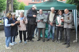 Theresa sowie 6 Besucher:innen und ein Ehrenamtlicher stehen vor einem grünen Pavillon und schauen zusammen auf die mitgebrachten Smartphones und Tablets.