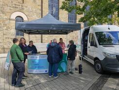 Mehrere Personen stehen um einen Pavillon herum, der neben dem Infomobil auf einem Marktplatz aufgestellt ist.