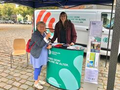 Petra (rechts) steht mit der Seniorin Karola (links) unter einem Pavillon am Info-Stand. Karola steht ihren rechten Daumen nach oben und strahlt. 