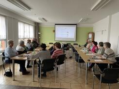 Die Teilnehmenden sitzen an zwei längeren Gruppentischen und schauen auf die Leinwand, auf der die Stationen des Digitalen Engels angezeigt werden. Die Wand hinter der Leinwand ist hellgrün.