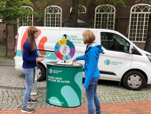 Petra steht mit Melanie Sing von der Stadt Elmshorn am Glücksrad vor dem Info-Mobil. Frau Sing dreht das Glücksrad und Petra hält Zettel mit Fragen in der Hand.