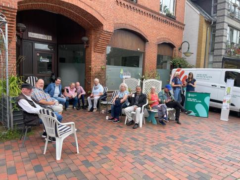 Das Info-Mobil steht vor dem Café. Petra bespricht mit einer Seniorin ein individuelles Thema. Daneben sitzen viele Senior:innen auf Stühlen und trinken Kaffee.