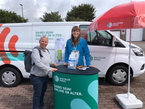 Frau Düßmann und Petra stehen am Info-Stand vor dem Info-Mobil und lächeln in die Kamera.