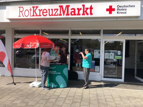 Petra steht gemeinsam mit einer Dame am Info-Stand vor dem RotKreuzMarkt. Eine weitere Dame läuft zum Stand.