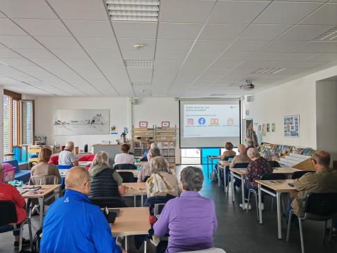 Die Teilnehmenden sitzen an Tischen im Raum und blicken zur Präsentation an der Leinwand