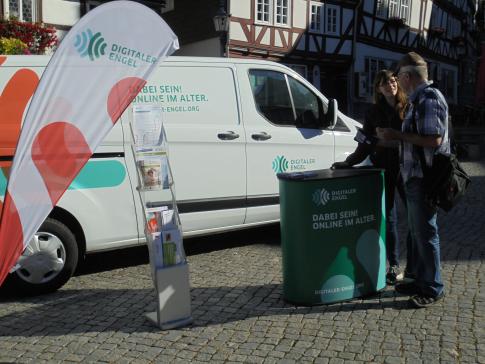 Petra steht mit dem Senioren Achim am Info-Stand des Digitalen Engel. Die beiden unterhalten sich.