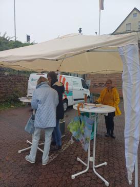 Petra steht mit einer Dame am Info-Stand. Der Info-Stand ist mit einem Pavillon vor dem Regen geschützt.