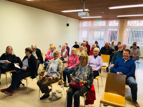 Knapp 30 Senior:innen sitzen auf Stühlen in fünf Reihen ohne Tische. Einige gucken in die Kamera - andere gucken weg.