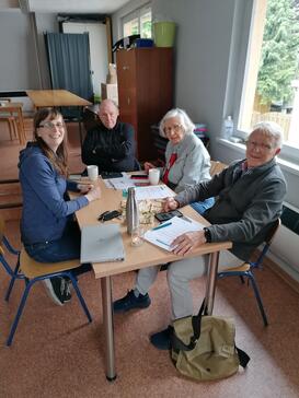 Petra sitzt mit drei Senioren und einer Seniorin an einem Tisch. Alle vier gucken nach vorne in die Kamera.
