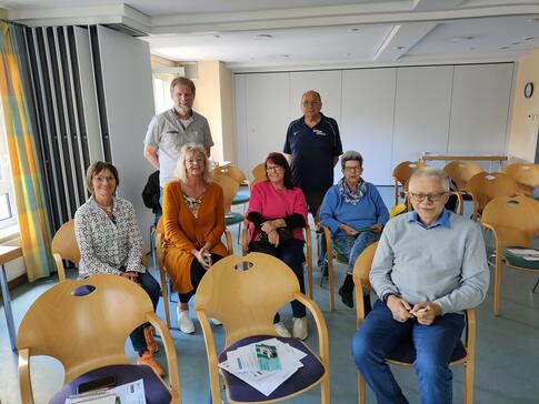 Auf dem Gruppenfoto sitzen fünf Frauen und ein Mann auf Stühlen. Hinter ihnen stehen zwei Männer. Alle sieben schauen in die Kamera und lächeln.