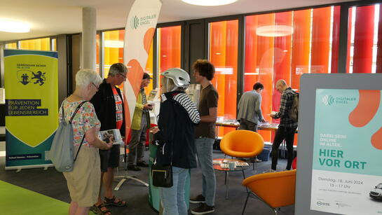 Besucher:innen stehen im Foyer der Bücherei vor dem Infostand und unterhalten sich miteinander und blicken zu Johannes 