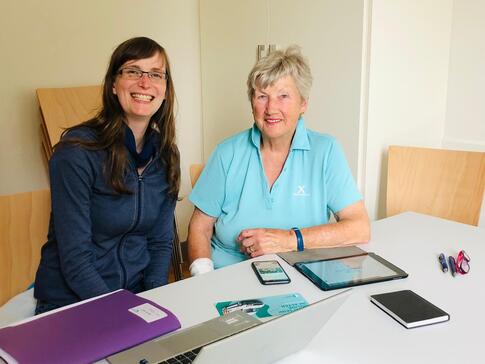 Petra sitzt mit einer Seniorin an einem Tisch. Vor ihnen liegen zwei Smartphones und ein Tablet.