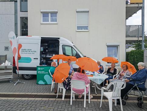 Mehrere Damen und Herren sitzen draußen vor dem Infomobil in einem Kreis um einen Tisch herum und halten Regenschirme in den Händen