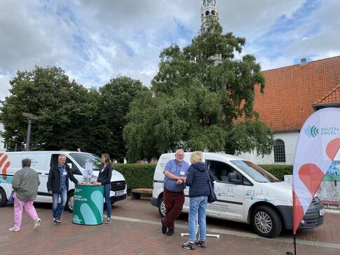 Das Info-Mobil (links) steht neben dem Mobil vom offenen Kanal Westküste (rechts). Vor dem rechten Mobil steht Andreas mit einer Dame. Links stehen Horst und Petra am Info-Stand. Eine Seniorin verlässt gerade den Stand.