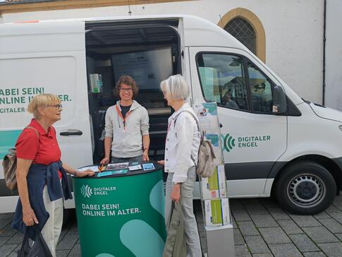 Zwei Damen stehen neben Johannes vor dem Infomobil und unterhalten sich