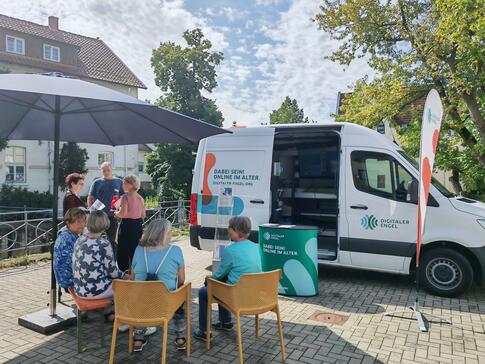 Mehrere Besucherinnen sitzen auf Stühlen vor dem Infomobil und unterhalten sich, außerdem steht Frau Röthe mit einer weiteren Besucherin und einem Besucher an einem Stehtisch