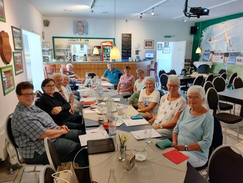 Zwölf Teilnehmende (neun Frauen und drei Männer) sitzen an einem langem Tisch im Vereinsheim und gucken nach vorne in die Kamera.