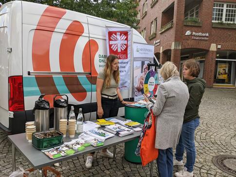 Theresa und eine Besucherin, die links neben ihr steht, sind von hinten zu sehen und hören einer Caritas-Mitarbeiterin zu, die am gemeinsamen Stand vor dem Infomobil steht.