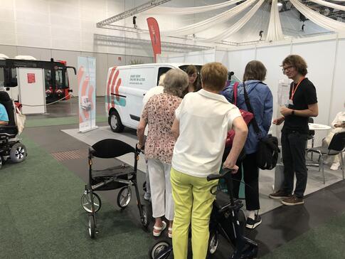 Johannes und Petra beraten vier Seniorinnen am Infostand, zwei Seniorinnen sind mit Rollator gekommen. Links im Hintergrund ist das Infomobil zu sehen.