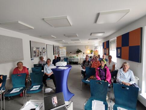 Auf der linken und rechte Seite des Veranstaltungsraumen sitzen elf Teilnehmende sowie die Ansprechpartnerinnen des Senioren- und Pflegestützpunkt. In der Mitte des Raumes steht ein Beamer und ganz vorne im Bild ist ein Laptop zu sehen. 