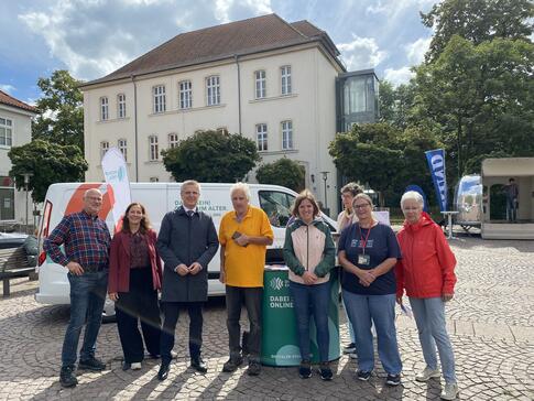 Theresa, die Projektleitung Katharina Kunze, der Abteilungsleiter des BMFSFJ sowie fünf Besucher:innen aus Lauenburg stehen auf einem öffentlichen Platz vor dem Infomobil und lächeln in die Kamera.