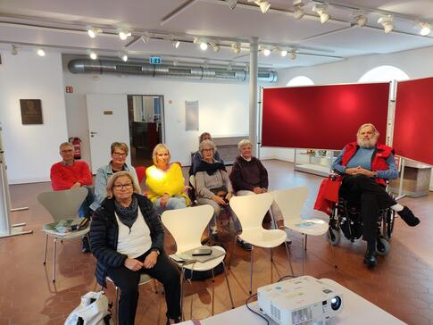 In einem Raum des Museums "zeiTTor" sitzen sieben Personen (ein Mann und sechs Frauen) in drei Stuhlreihen. Am rechten Bildrand sitz ein weiterer Mann im Rollstuhl. 