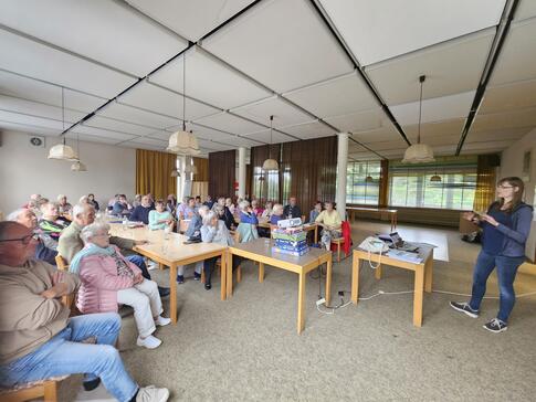 Auf der linken Seite des Bildes sitzen 45 Seniorinnen und Senioren auf Stühlen an  Tischen. In der Mitte des Bildes steht ein Beamer auf Spiele-Packungen. Rechts im Bild steht Petra mit ihrem Laptop und erklärt etwas. 