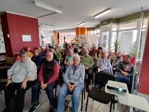 Über dreißig Senior:innen sitzen an über fünf Stuhlraumen in der Kleinen Markthalle in Stendal und blicken nach vorne.