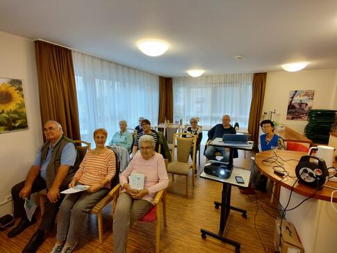 In einem Raum sitzen neun Seniorinnen und Senioren auf Stühlen ohne Tische. Rechts im Bild stehen ein Beamer und ein Laptop.