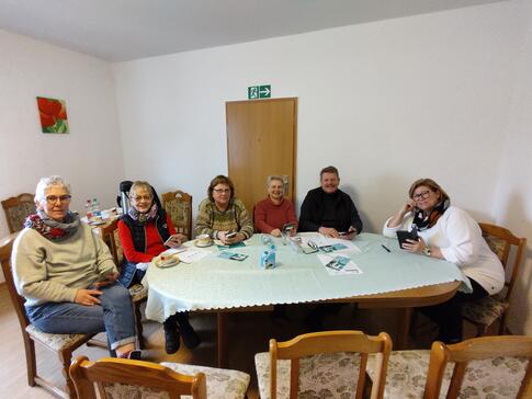 Fünf Seniorinnen und ein Senior sitzen an einem ovalen Tisch und lächeln vorne in die Kamera.