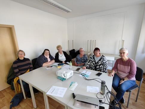 Sechs Frauen sitzen an zwei zusammengestellten Tischen und gucken in die Kamera. Auf den Tischen liegen ein Beamer, ein Laptop und Unterlagen.