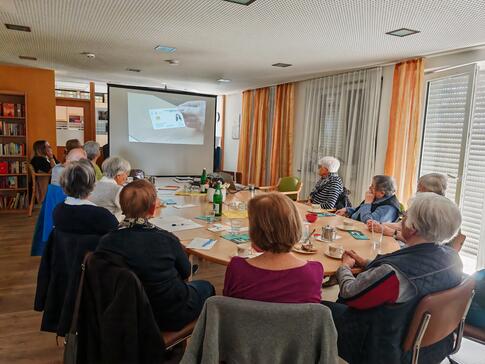 Mehrere Personen sitzen an einem runden Tisch und blicken nach vorne zur Leinwand auf der ein Video abgespielt wird