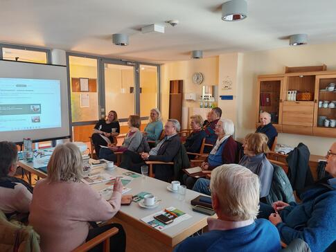 Die Teilnehmenden sitzen auf Stühlen um einen großen Tisch herum und blicken nach vorne auf eine Leinwand