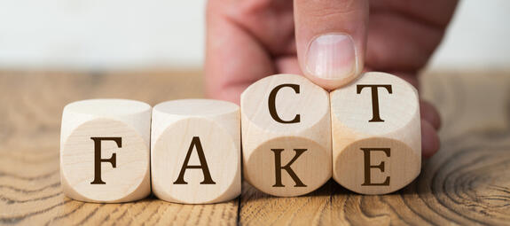 Das Bild zeigt Würfel auf einem Holztisch, die die Wörter "Fake" und "Fact" bilden. Eine Hand kommt von oben und dreht die Würfel, sodass aus "Fact" das Wort "Fake" wird. 