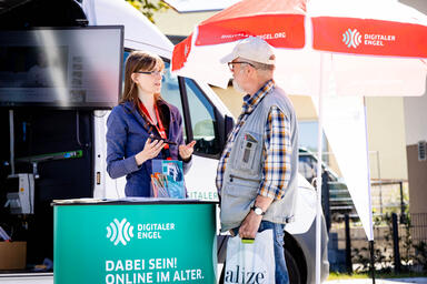 Zwei Menschen vor einem Infomobil unterhalten sich, Frau hält Smartphone in der Hand